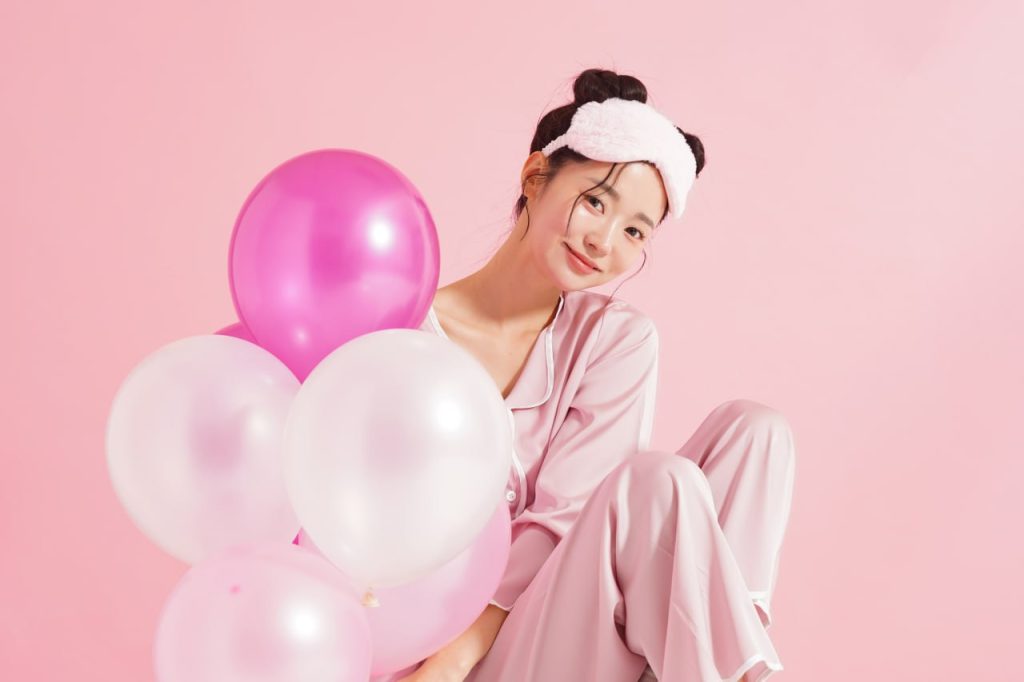 Young woman in pink satin pajamas with fluffy headband holding pink and white balloons for Overnight Miracle skincare