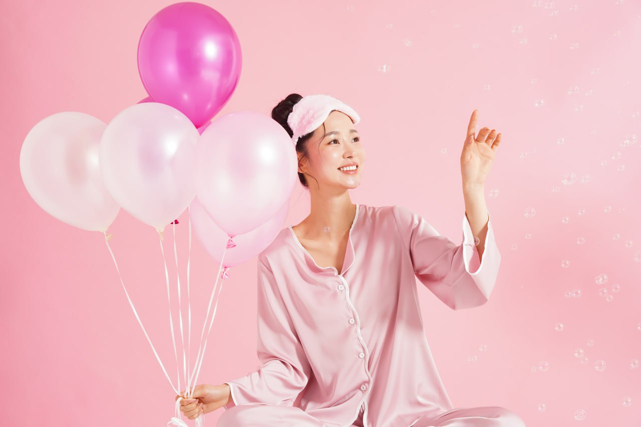 Smiling woman in pink satin pajamas and eye mask holding pink balloons against a pink backdrop for Overnight Miracle skincare