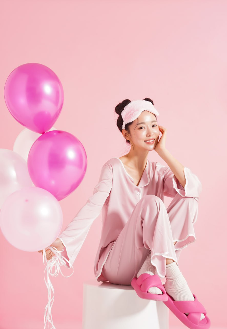 Young woman in pink sleepwear and eye mask holding pink and white balloons, smiling to showcase overnight skin glow treatment