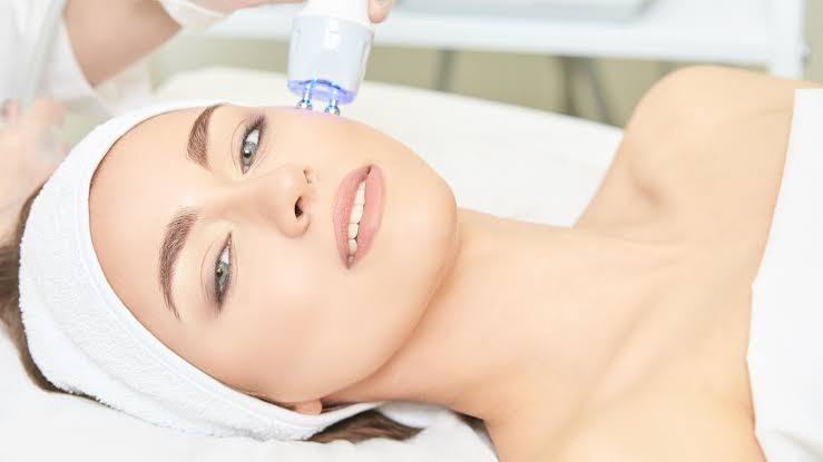 Woman receiving blue-light skin rejuvenation facial treatment at clinic, lying with headband and relaxed expression