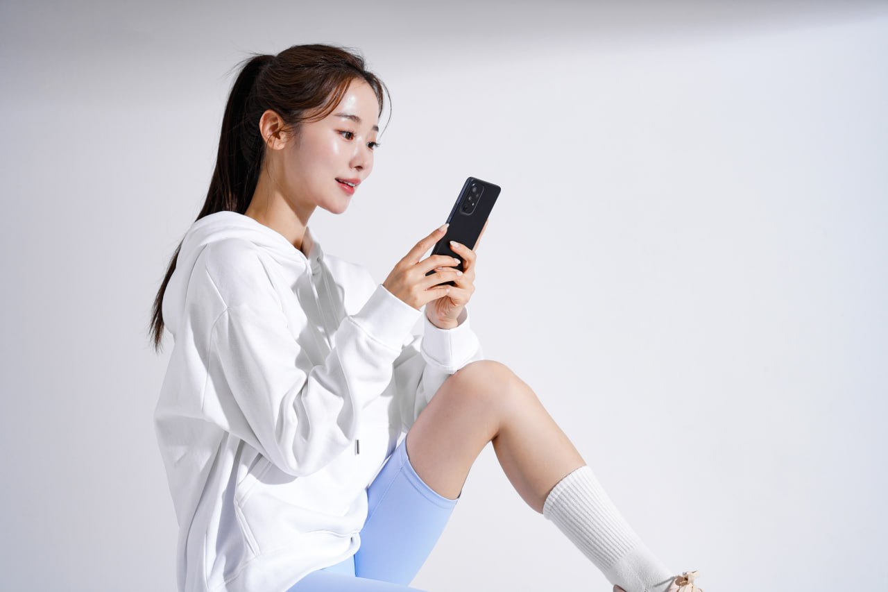 Young woman in white hoodie and blue shorts checking her phone after a workout, showing clear hydrated skin, sporty look