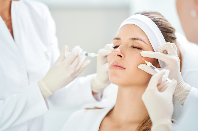 Woman receiving Botox injections to cheeks by gloved clinicians in a bright dermatology clinic treatment room