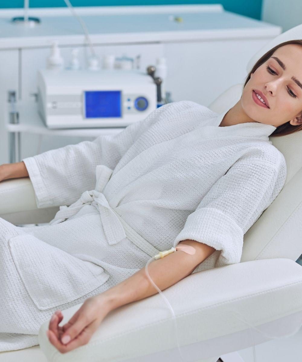 Woman in a white spa robe reclining with an IV vitamin drip in a dermatology clinic treatment chair, relaxed expression