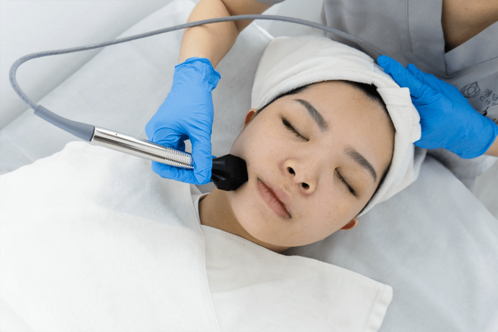 Patient lying with eyes closed as clinician in blue gloves performs Korean V-Lifting HIFU on lower face and jawline