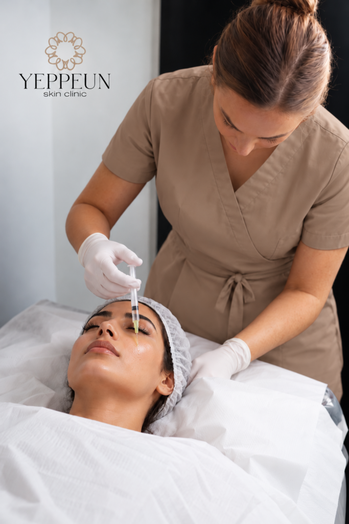 A female patient lies on a treatment bed with her eyes closed as a skin expert holds a syringe of light yellow serum near her under-eye area.